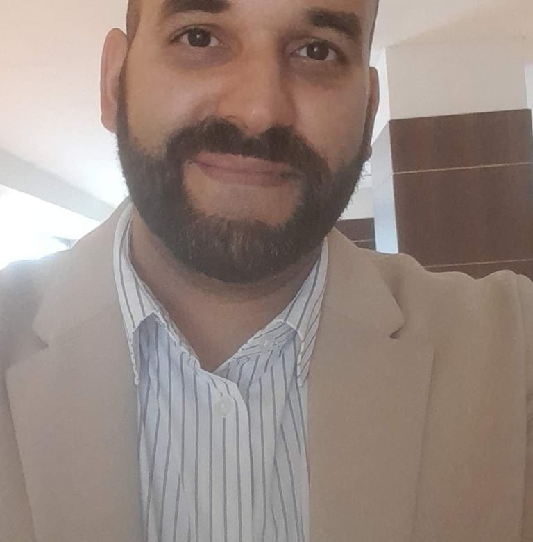 A man with a full beard wearing a beige blazer and light blue striped shirt